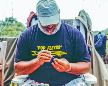 Man sitting outdoors, wearing a cap and a "Pop Fleyes" t-shirt, focused on tying a fishing fly.