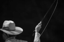 Man in a hat casting a fishing rod against a dark background in black and white.