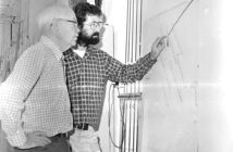 Two men in plaid shirts study a wall-mounted diagram, one pointing at it, in a narrow corridor.