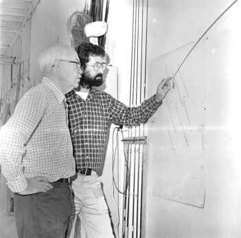 Two men in plaid shirts study a wall-mounted diagram, one pointing at it, in a narrow corridor.