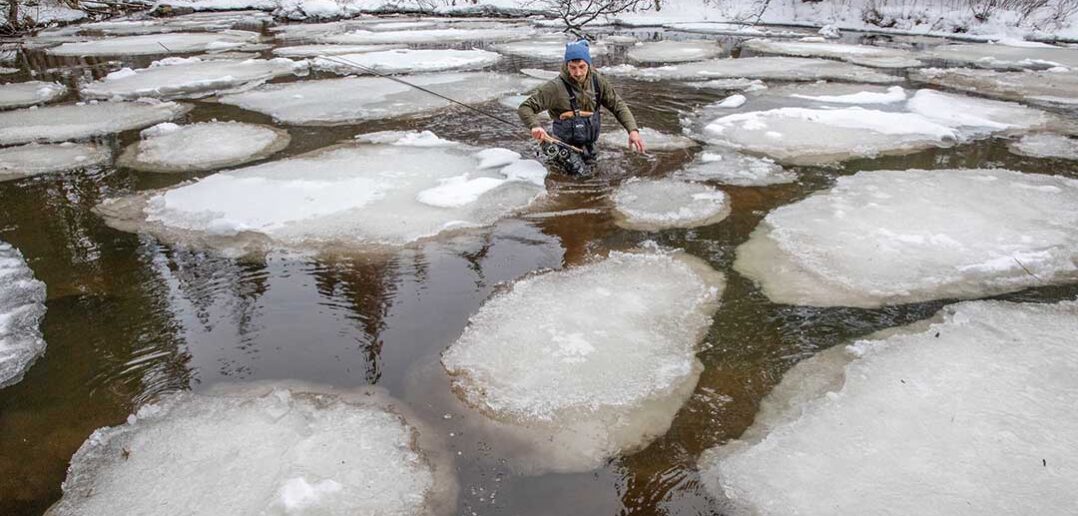 Person wading through an icy river surrounded by large ice patches on a winter day.