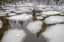 Person wading through an icy river surrounded by large ice patches on a winter day.