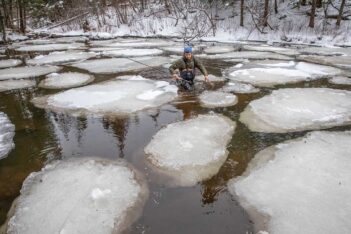 Person wading through an icy river surrounded by large ice patches on a winter day.