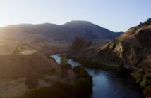 Sun setting over a winding river with hills and sparse vegetation, casting long shadows in a tranquil landscape.