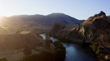 Sun setting over a winding river with hills and sparse vegetation, casting long shadows in a tranquil landscape.