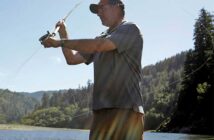 A man wearing a cap and short-sleeved shirt is fly-fishing in a scenic river with a forested hillside in the background. Sunlight streams across the scene.