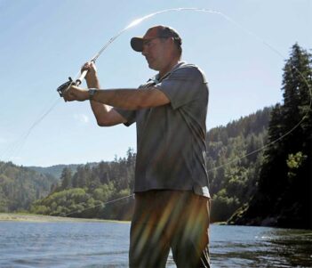 A man wearing a cap and short-sleeved shirt is fly-fishing in a scenic river with a forested hillside in the background. Sunlight streams across the scene.