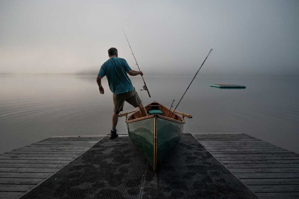 A person stands on a dock with a fishing rod and a canoe, facing a foggy lake.