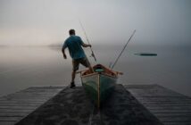 A person stands on a dock with a fishing rod and a canoe, facing a foggy lake.