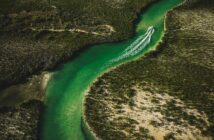 Aerial view of a green river winding through dense greenery, with a boat creating a wake as it travels upstream.