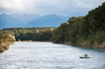 Small boat on a wide river with two people, surrounded by lush green forest and distant mountain range under a cloudy sky.