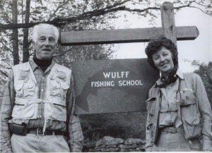 Two people stand in front of a sign for "Wulff Fishing School." They are both wearing outdoor vests. Trees are in the background.