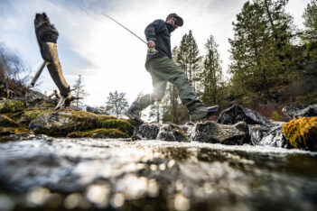 Person wearing outdoor gear and holding a fishing rod walks across rocks in a stream surrounded by trees, with sunlight shining in the background.