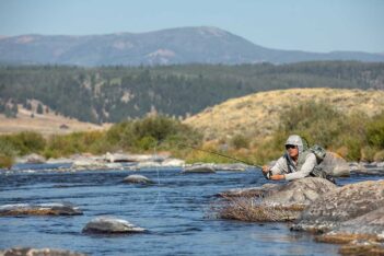 Person fly fishing on a rocky riverbed, surrounded by grassy banks and distant mountains under a clear blue sky.