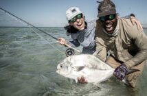 Two people smiling widely hold a large fish in shallow water. One person wears a cap and sunglasses, the other wears a hat and sunglasses.