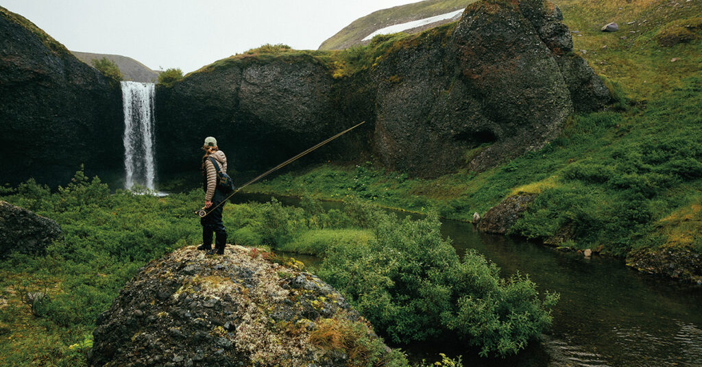 A person stands on a rock holding a fishing rod, overlooking a river and waterfall surrounded by green hills and vegetation.