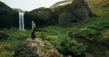 A person stands on a rock holding a fishing rod, overlooking a river and waterfall surrounded by green hills and vegetation.