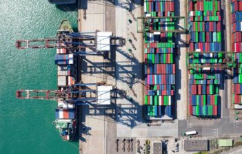 Aerial view of a busy shipping port with cranes, stacked multicolored cargo containers, and a docked cargo ship next to turquoise water.